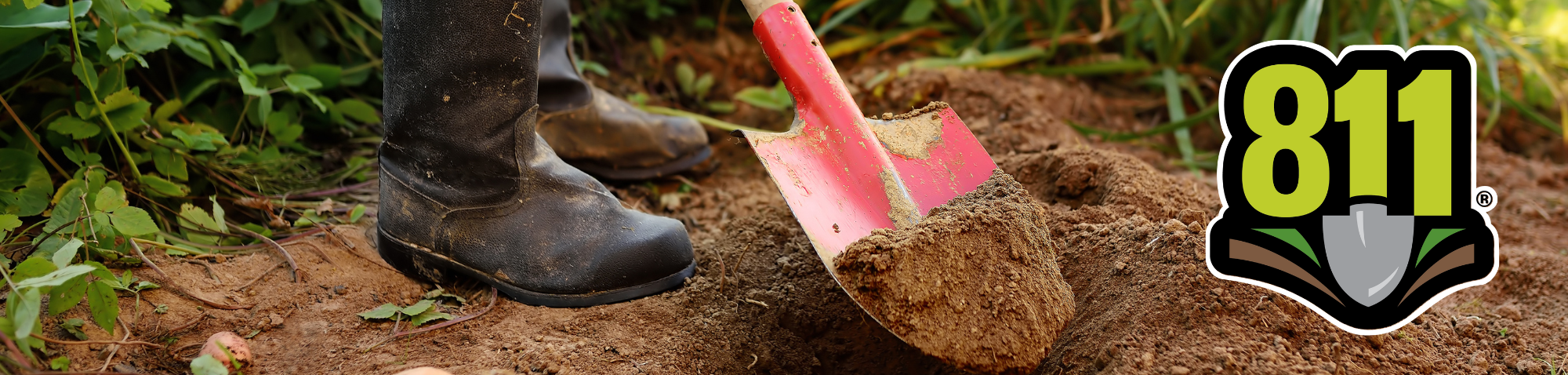 Black rubber rainboots standing on a patch of dirt. A red shovel full of dirt is above a hole in the ground.